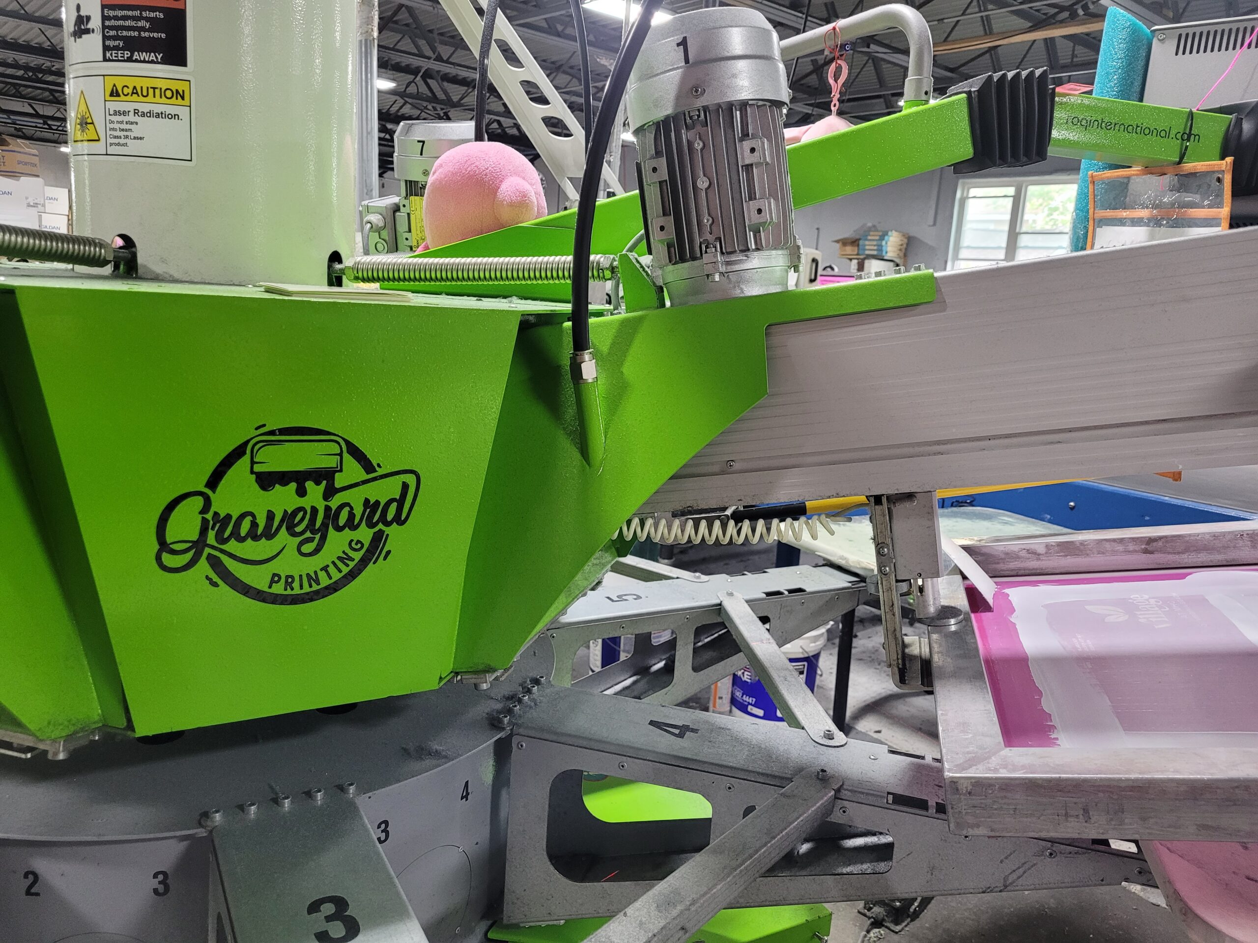 A green screen-printing machine with Graveyard Printing on the side is in operation. The image shows a light industrial setting with printing equipment, including a pink screen frame ready for use.