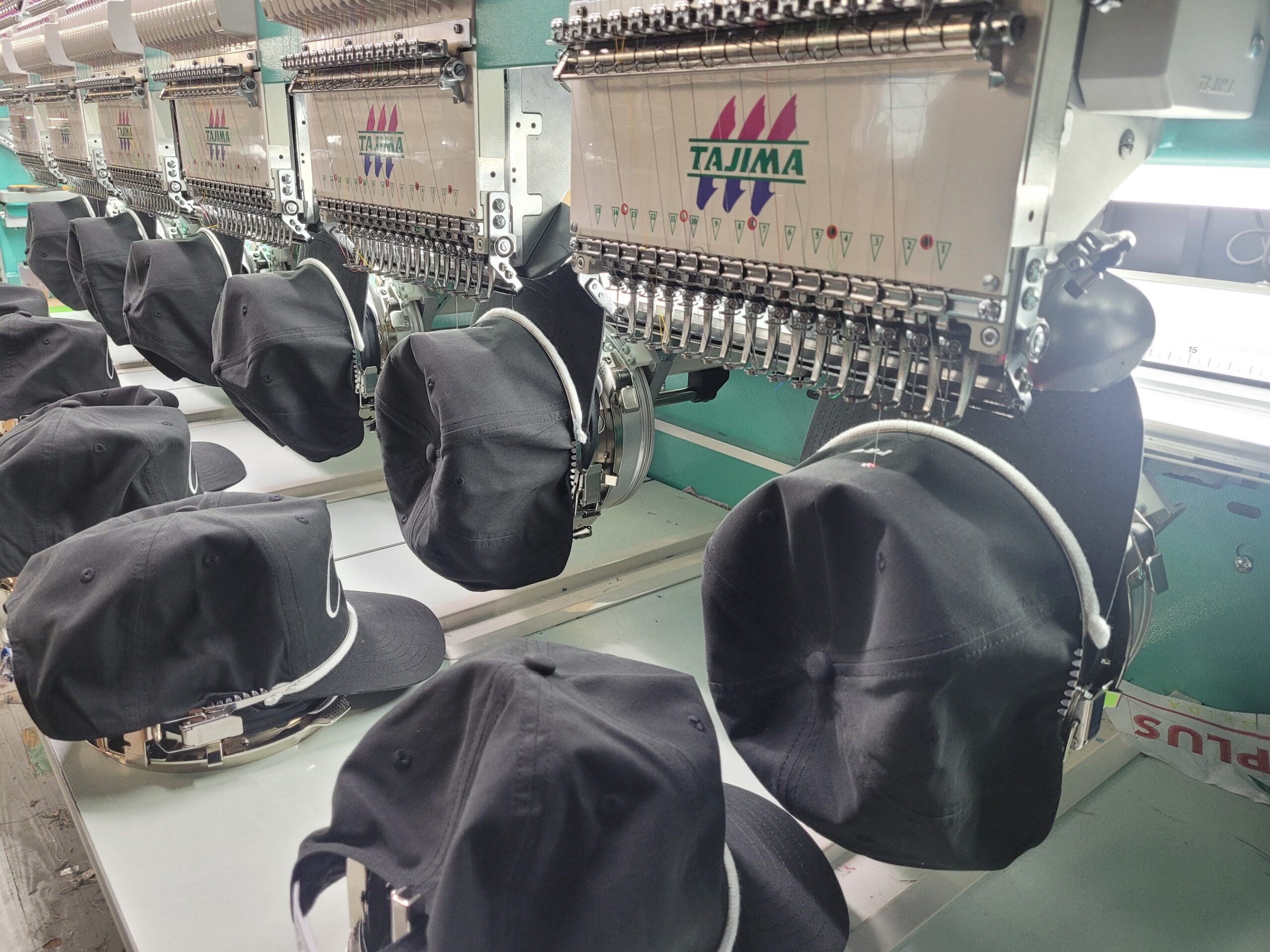 Show thumbnail preview Black baseball caps being embroidered on a row of industrial Tajima embroidery machines in a factory setting.