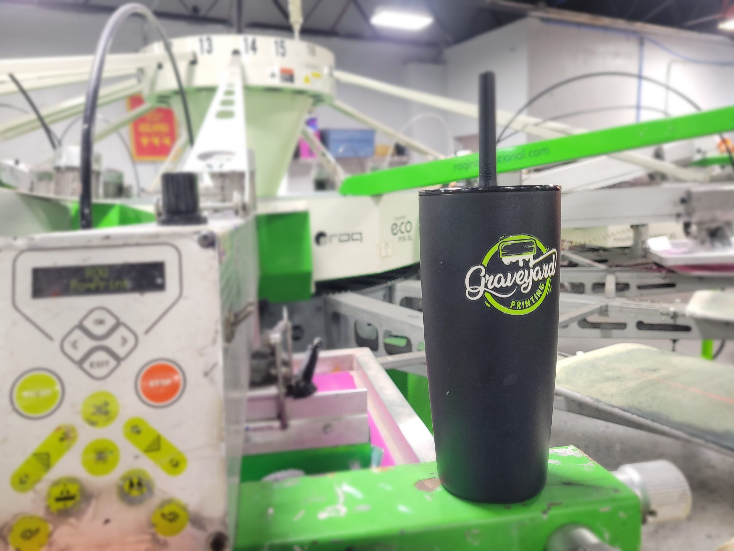 A black tumbler with the text Graveyard Printing is placed on a green surface. In the background, there are screen printing machines in an industrial setting. The area is well-lit, showcasing equipment and printed designs.