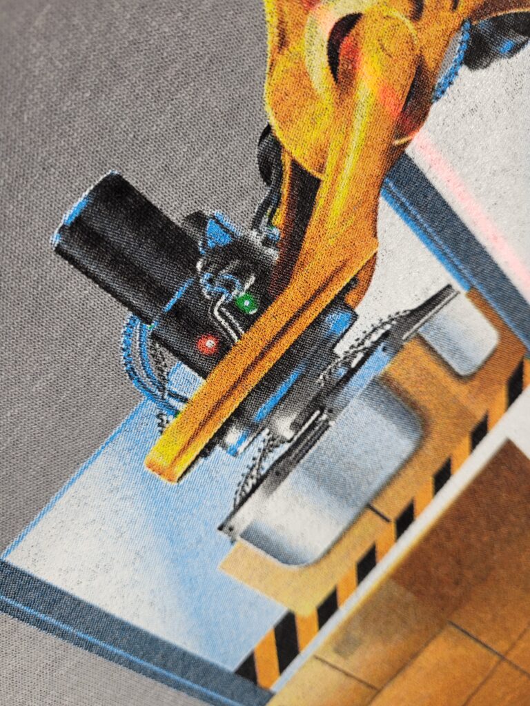 A robotic arm in a factory setting, holding a metallic object. The arm is yellow with black and red components. The background features industrial elements like a conveyor belt and yellow and black caution markings.