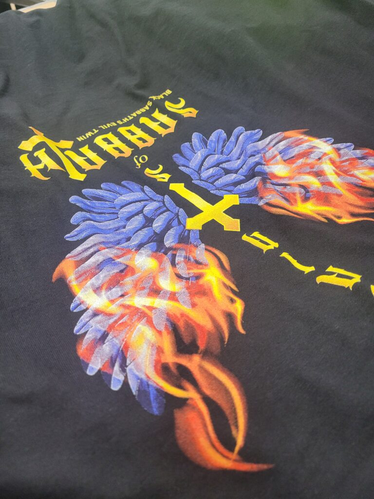 A black T-shirt with a vibrant design featuring blue angel wings intertwined with fiery elements. The text above the wings reads All the Saints in yellow Gothic-style font.