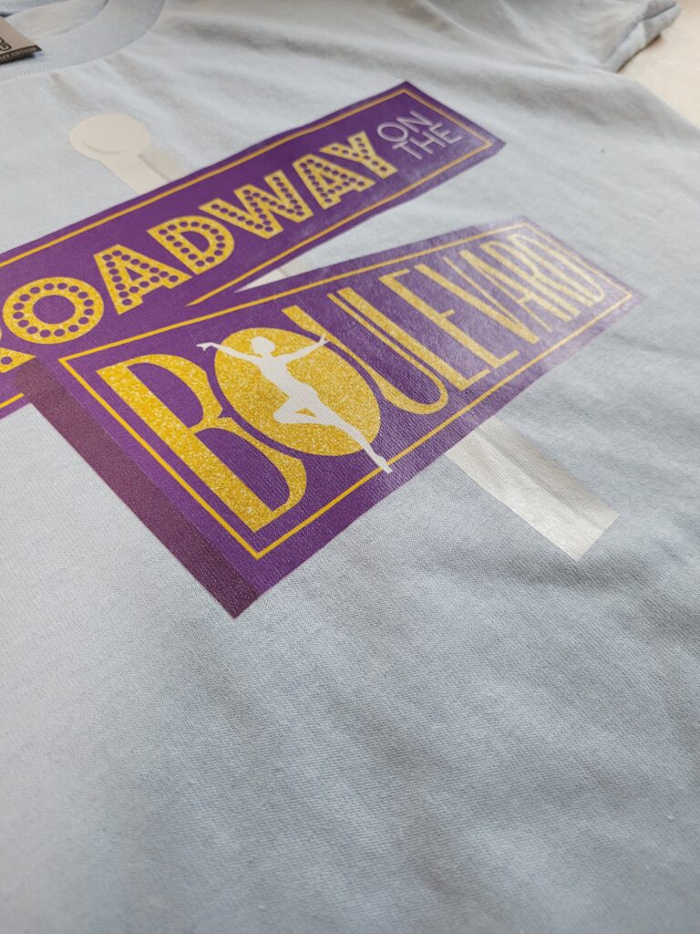 A light gray T-shirt with a colorful print design. The text reads Broadway on the Boulevard in bold letters, featuring a silhouette of a dancer. The colors include purple, gold, and white, creating a vibrant and theatrical theme.