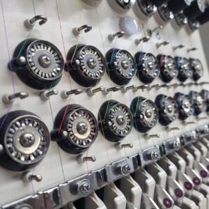 Show thumbnail preview Close-up of a mechanical knitting machine, showing a series of circular tension discs with colorful threads passing through them. Metal hooks and levers are visible below, part of the intricate machinery.