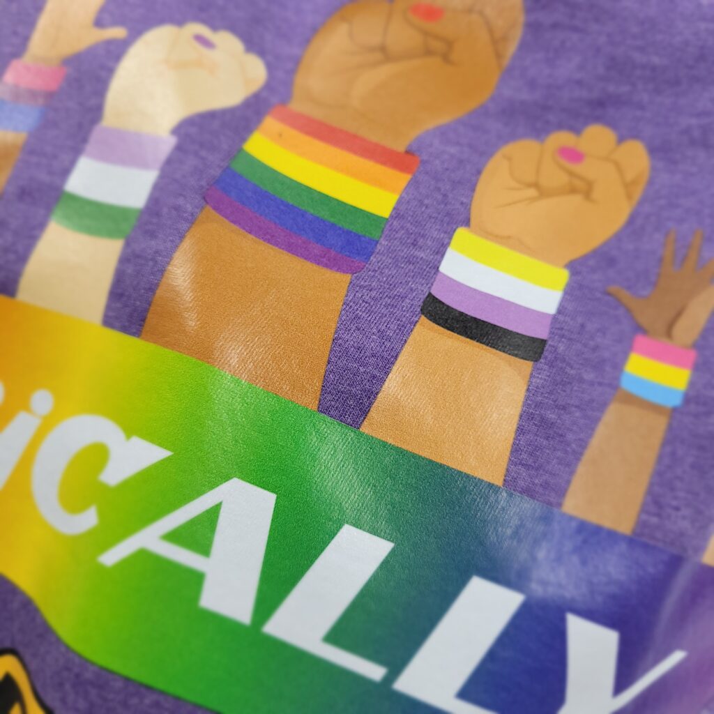 Illustration of raised fists with diverse skin tones, each wearing a rainbow wristband. The background features the word ALLY partially visible on a rainbow gradient banner.