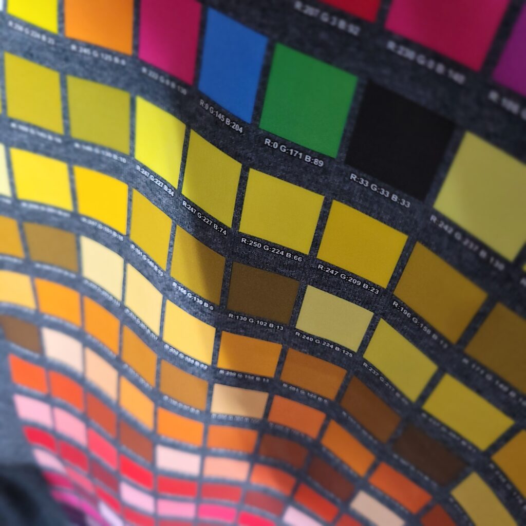 A fabric swatch book displaying numerous color samples arranged in rows and columns. The swatches range from yellows and oranges to greens and blues, each labeled with color codes. The fabric appears wavy with varying light reflections.